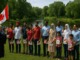 A diverse group of immigrants stands outdoors in a line, raising their right hands as they take the Canadian Oath of Citizenship in front of an officiant. They hold small Canadian flags and certificates, with a scenic park, pond, and trees in the background. The ceremony takes place on a sunny day in Winnipeg’s Assiniboine Park.